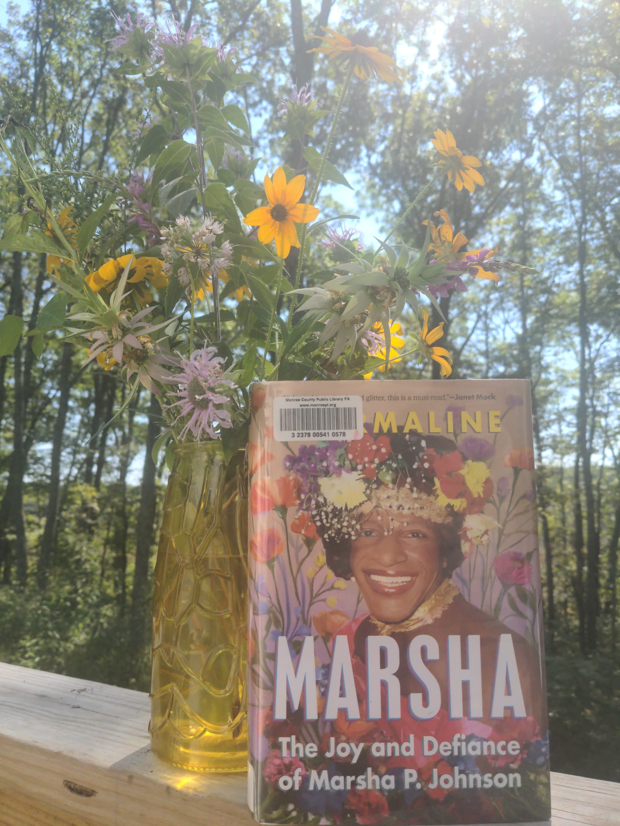 A picture of the book Marsha by Tourmaline, propped up beside a golden vase of wildflowers, a wooded backyard behind it. The cover features a gorgeous photograph of Marsha P. Johnson with flowers in her hair and more flowers illustrated all around, a glowing smile on her face.
